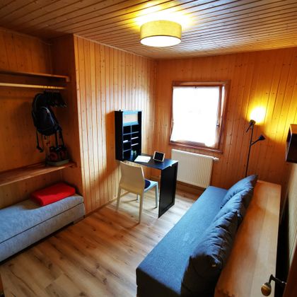Ein kleines Zimmer mit Holzverkleidung. Es gibt ein Bett, einen Schreibtisch und einen Stuhl, sowie ein Fenster, das natürliches Licht hereinlässt.