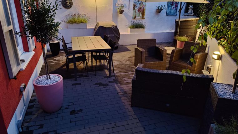 Ein gemütlicher Patio mit einem großen Tisch, Stühlen und einem Sonnenschirm. Die Umgebung ist gut beleuchtet und es gibt einige Pflanzen und Dekorationen an der Wand.