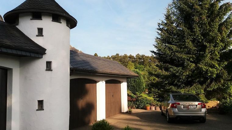Ein elegantes Haus mit einem runden Turm und einem grauen Auto in der Einfahrt. Umgeben von Bäumen und grüner Landschaft.