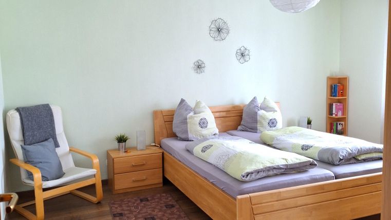 Ein helles Schlafzimmer mit zwei Betten und gemütlicher Ausstattung. Es gibt einen bequemen Stuhl und Bücherregale an der Wand.