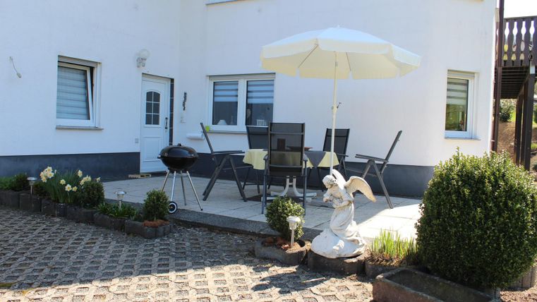 Ein gemütlicher Außenbereich mit einem Tisch, Stühlen und einem Sonnenschirm. Es gibt einen Grill und einige Pflanzen sowie eine Statue.