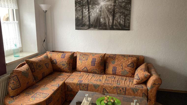 Ein gemütliches Wohnzimmer mit einer orangefarbenen Sofa-Ecke und einem Glastisch. An der Wand hängt ein Schwarz-Weiß-Bild eines Waldes.