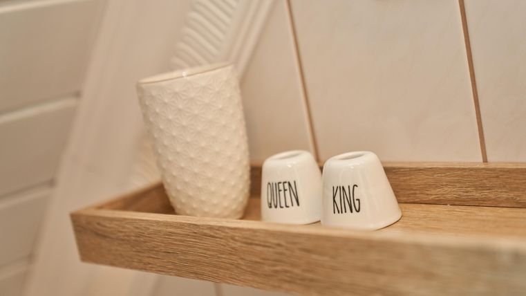 Eine dekorative Ablage mit einem weißen Becher und zwei kleinen Tassen, die mit "Queen" und "King" beschriftet sind. Die Umgebung hat ein warmes, gemütliches Ambiente.