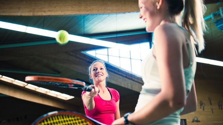 Zwei Frauen spielen Tennis in einer Sporthalle. Eine Spielerin ist bereit, den Ball zu treffen, während die andere lächelt.