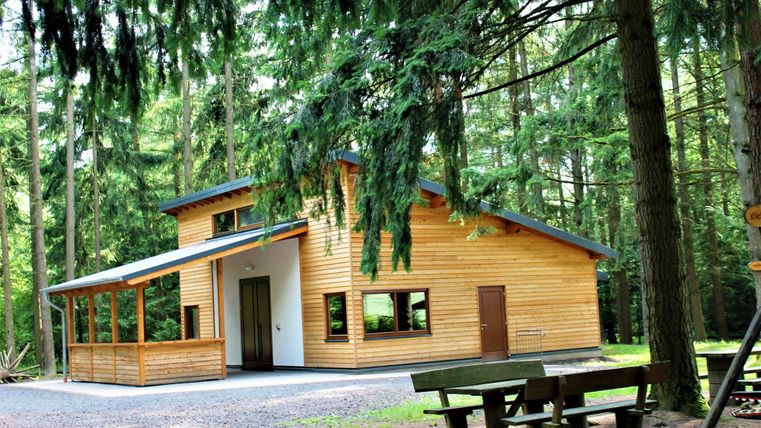 Ein modernes Holzhaus in einem Waldgebiet. Vor dem Haus stehen Bänke und Tische, umgeben von hohen Bäumen.