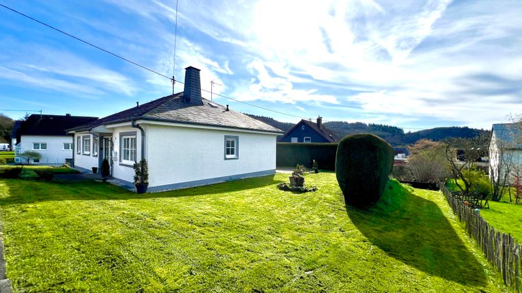 Ein kleines, weißes Haus mit einem gepflegten Garten. Im Hintergrund sind sanfte Hügel und ein blauer Himmel mit wenigen Wolken zu sehen.