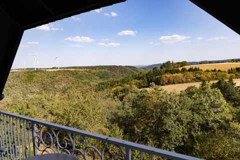 A picturesque view over green hills and forests. In the background, wind turbines and a clear sky can be seen.