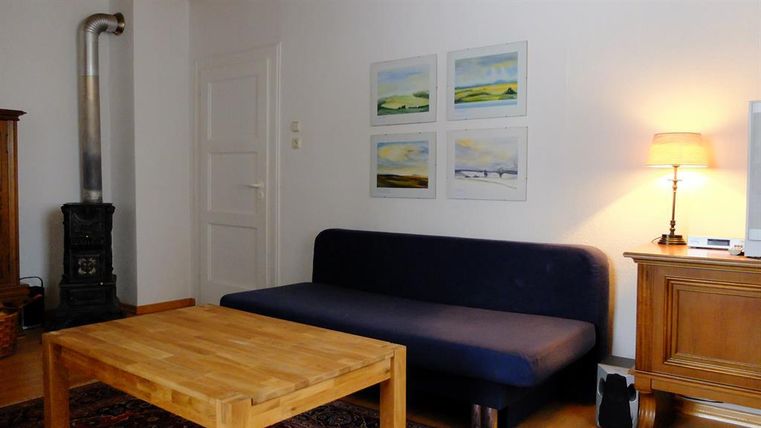 Ein einfaches Wohnzimmer mit einem blauen Sofa und einem Holzkaffee Tisch. An der Wand hängen mehrere Bilder und eine Lampe sorgt für warmes Licht.