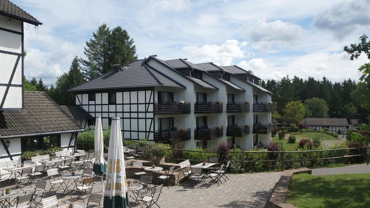 Ein malerisches Hotel mit traditioneller Architektur und modernen Balkonen. Im Vordergrund befinden sich Tisch und Stühle auf einer Terrasse, umgeben von grünem Garten.