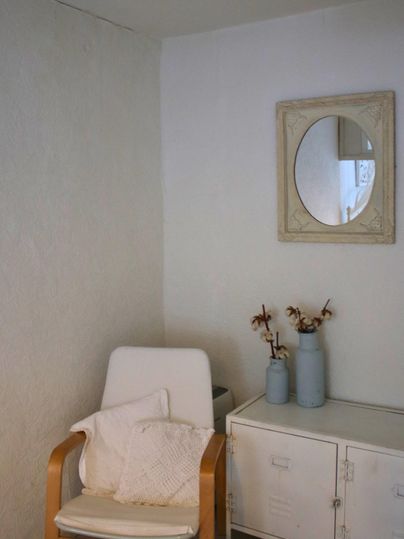 Ein heller Raum mit einem bequemen Stuhl und einem kleinen Tisch. Auf dem Tisch stehen zwei Vasen mit Trockenblumen und ein runder Spiegel hängt an der Wand.