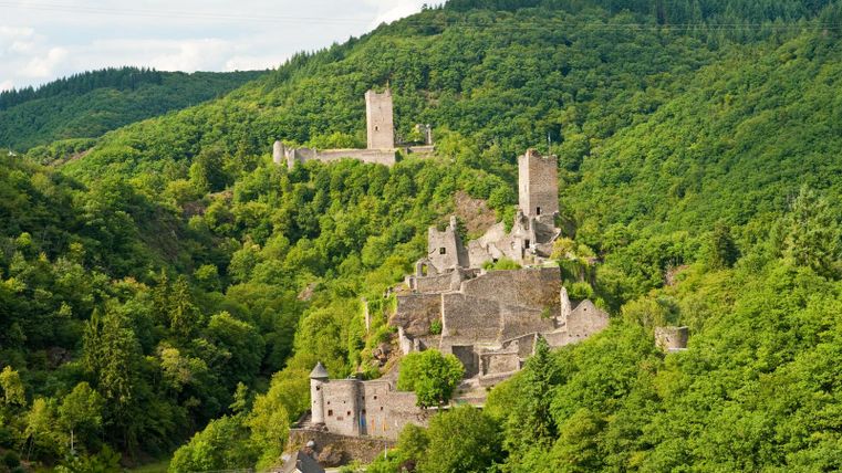 Een schilderachtig landschap met indrukwekkende kasteelruïnes, omringd door groene heuvels. Op de achtergrond zijn andere kastelen zichtbaar die de historische sfeer versterken.