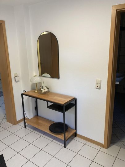 Ein moderner Flur mit einem stilvollen Tisch und einem runden Spiegel. Die Wände sind weiß, und der Boden ist mit Fliesen bedeckt.