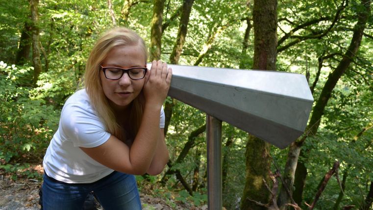Eine Frau lehnt sich an ein Fernrohr in einem Wald. Die Umgebung ist grün und von Bäumen umgeben.