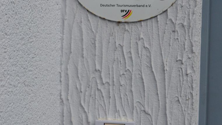 Ein Schild mit der Aufschrift "Ferienhaus/Ferienwohnung" hängt an einer weißen Wand. Darunter befindet sich ein Klingelschild.