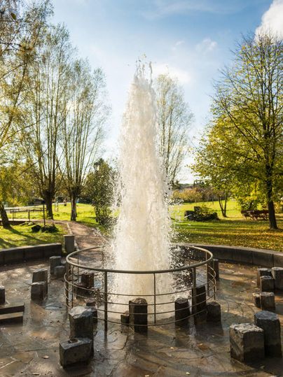 Ein Springbrunnen sprudelt in einem Park, umgeben von Bäumen und grünem Gras. Der blaue Himmel und das Sonnenlicht schaffen eine entspannte Atmosphäre.