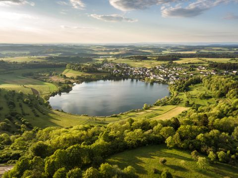 Ein malerischer See umgeben von grünen Wäldern und sanften Hügeln. Im Hintergrund ist ein kleines Dorf zu sehen.