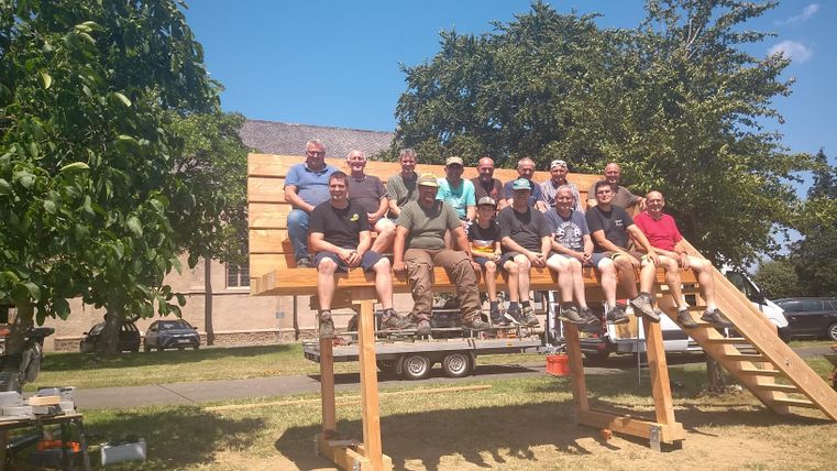 Eine Gruppe von Männern sitzt auf einer großen Holzplattform im Freien. Es ist ein sonniger Tag mit blauem Himmel und einigen Bäumen im Hintergrund.