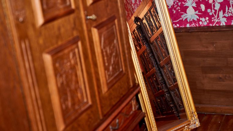 Ein großer, goldener Spiegel steht an einer Wand neben einem Holzschrank. Der Hintergrund ist mit floralem Muster in kräftigen Farben dekoriert.