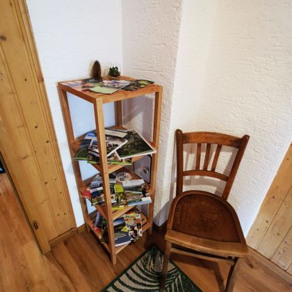 Ein Holzregal steht in der Ecke mit Postkarten und Zeitschriften. Daneben steht ein einfacher Holzstuhl auf einem Teppich.