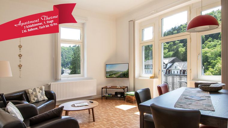 Ein modernes Wohnzimmer mit großen Fenstern, die auf eine grüne Landschaft blicken. Der Raum ist hell und freundlich mit einem gemütlichen Sofa und einem Esstisch.