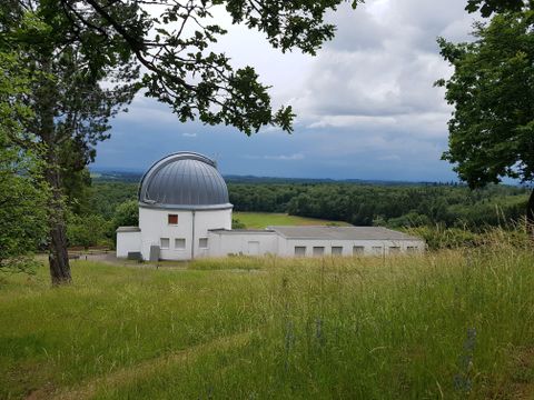 Eine Astronomie-Observatorium mit einer runden Kuppel, umgeben von grünen Wiesen und Bäumen. Der Himmel ist bewölkt und der Ausblick erstreckt sich über die Landschaft.