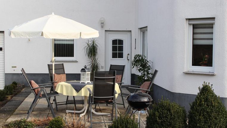 Ein gemütlicher Außenbereich mit einem Tisch und Stühlen, einem Sonnenschirm und einem Grill. Umgeben von kleinen Hecken und einer hellen Wand.