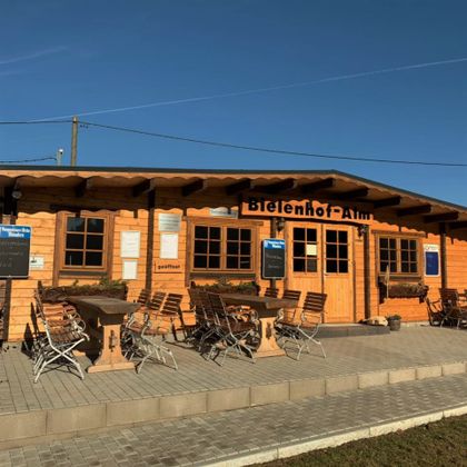 Holzgebäude der Bielenhof-Alm mit Tischen und Stühlen im Freien.