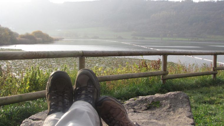 Eine friedliche Landschaft mit einem Fluss und sanften Hügeln im Hintergrund. Im Vordergrund liegen zwei Paar Füße auf einem Stein.