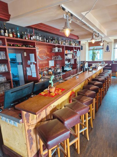 Eine gemütliche Bar mit einer langen Theke und Holzstühlen. Im Hintergrund stehen Regale mit verschiedenen Getränken.