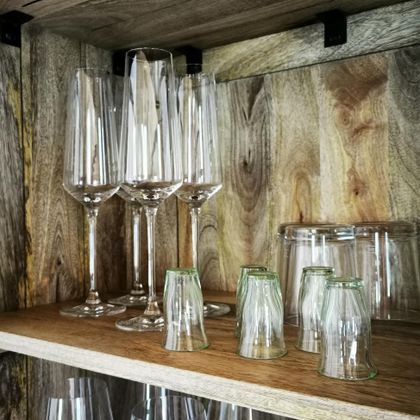 Ein Holzregal mit verschiedenen Gläsern. Oben stehen lange Sektgläser und unten kleine bunte Gläser.