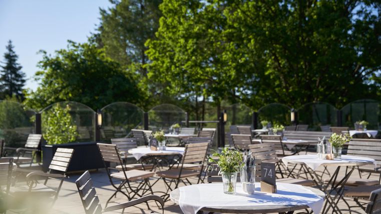 Eine ruhige Terrasse mit weißen Tischdecken und Holzmöbeln. Im Hintergrund grüne Bäume und blauer Himmel sorgen für eine angenehme Atmosphäre.