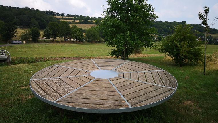 Ein runder Holztisch steht in einer grünen Wiese mit Bäumen im Hintergrund. Der Himmel ist leicht bewölkt, und die Landschaft wirkt ruhig und einladend.