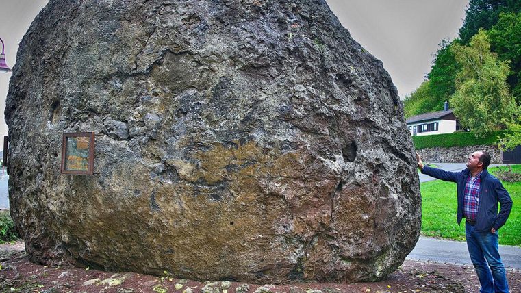 Ein großer Felsen steht an der Straße, neben einem kleinen Fenster. Ein Mann betrachtet den Felsen und berührt ihn.