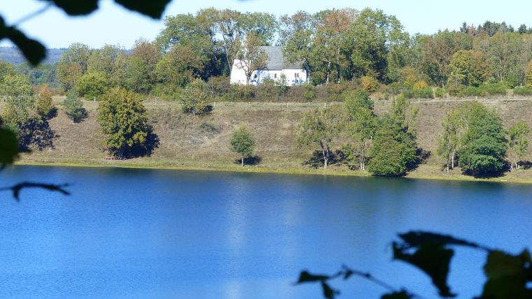 Ein ruhiger See mit klarem Wasser, umgeben von Bäumen. Im Hintergrund steht ein kleines, weißes Haus auf einem Hügel.