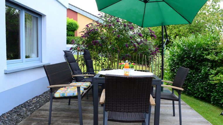 Eine gemütliche Terrasse mit einem Tisch und Stühlen. Ein grüner Sonnenschirm bietet Schatten und der Garten ist üppig bewachsen.