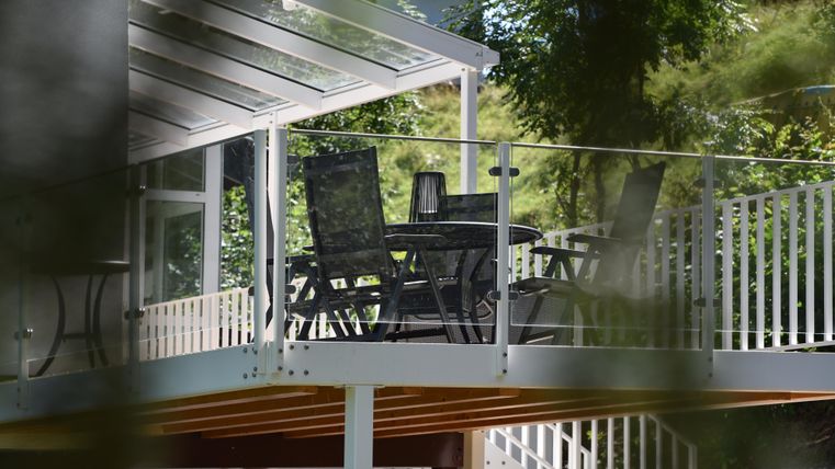 Eine moderne Terrasse mit einem Tisch und Stühlen, umgeben von Bäumen. Die Überdachung bietet Schatten und lädt zum Entspannen ein.