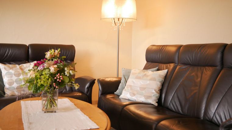 Ein gemütliches Wohnzimmer mit zwei schwarzen Ledersofas und einem runden Holztisch. Auf dem Tisch steht eine Vasen mit bunten Blumen und eine Tischlampe sorgt für warmes Licht.