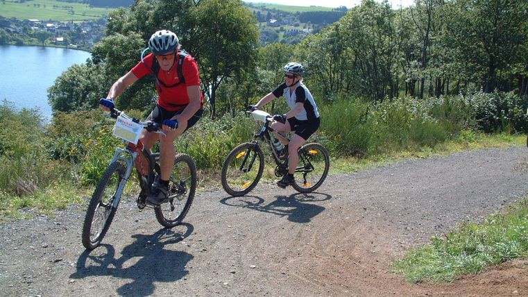 Zwei Mountainbiker fahren einen steilen Weg hinauf. Im Hintergrund ist ein schöner See und eine grüne Landschaft zu sehen.