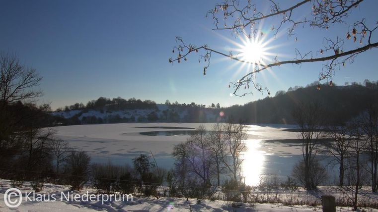 Eine winterliche Landschaft mit einem gefrorenen See und schneebedeckten Ufern. Die Sonne strahlt hell am Himmel und reflektiert im Wasser.