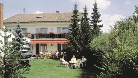 Ein gemütlicher Garten mit einer Gruppe von Personen, die an einem Tisch sitzen. Im Hintergrund steht ein Haus mit einem Balkon und grünen Bäumen.