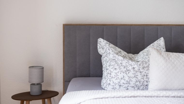 Ein modernes Schlafzimmer mit einem grauen Kopfteil und eleganten Kissen. Neben dem Bett steht ein kleiner Tisch mit einer Lampe.