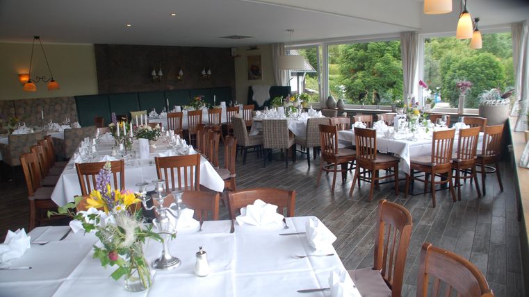 Ein helles Restaurant mit eleganten Holztischen und Stühlen. Die Tische sind ordentlich gedeckt und es gibt Fenster mit Blick auf die grüne Umgebung.