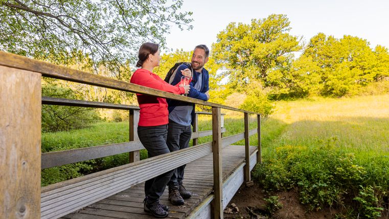 Zwei Personen stehen auf einer Holzbrücke in einer grünen Landschaft.