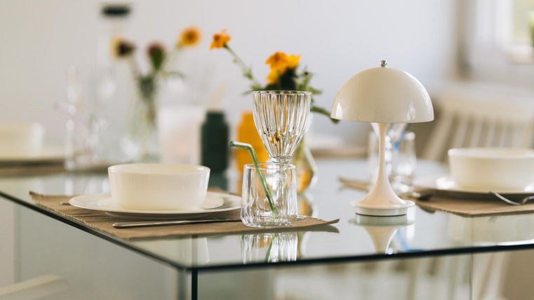 Ein eleganter Esstisch mit einem Glas und einer modernen Lampe. Auf dem Tisch stehen Blumen und Geschirr in einer hellen, einladenden Atmosphäre.