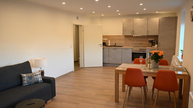 Eine gemütliche Wohnung mit einem modernen Wohnzimmer und einer offenen Küche. Der Essbereich ist mit einem Holztisch und orangenen Stühlen ausgestattet.