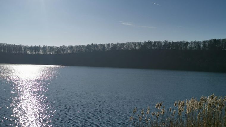 Ein ruhiger See unter einem klaren blauen Himmel. Die Sonne spiegelt sich im Wasser und umgeben ist der See von Bäumen.