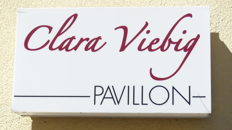 Ein Schild mit dem Namen "Clara Viebig Pavillon". Es hat eine einfache, elegante Schrift auf einem weißen Hintergrund.