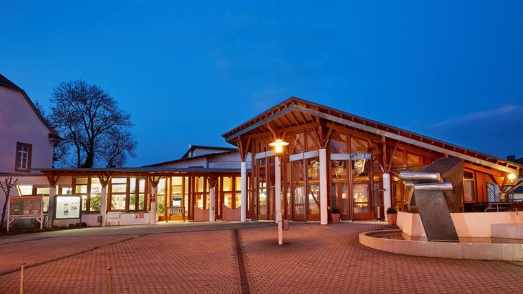 A modern building with wooden architecture and large windows. It is evening and the sky is blue, illuminated by lights in the entrance area.