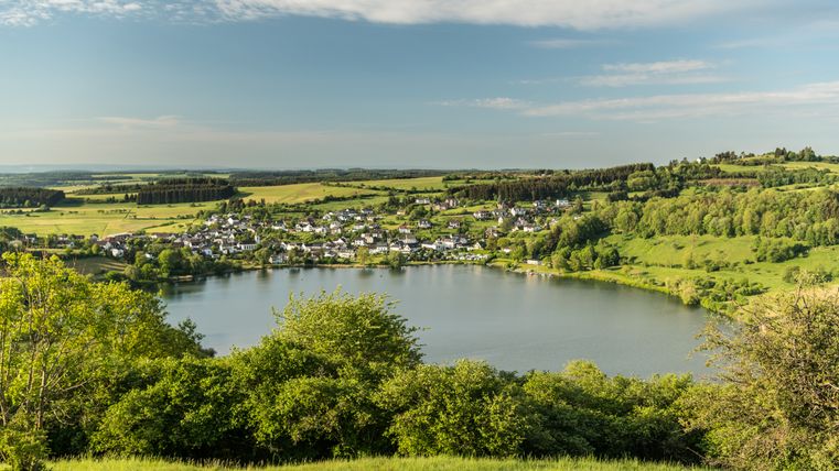 Blick auf das Schalkenmehrener Maar in der Eifel mit umliegendem Dorf und grüner Landschaft.