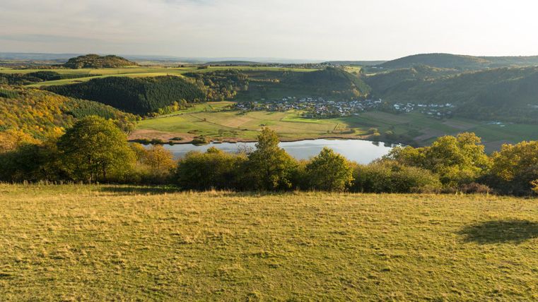 Eine malerische Landschaft mit sanften Hügeln und einem ruhigen See. Im Hintergrund sind kleine Dörfer und grüne Felder zu sehen.